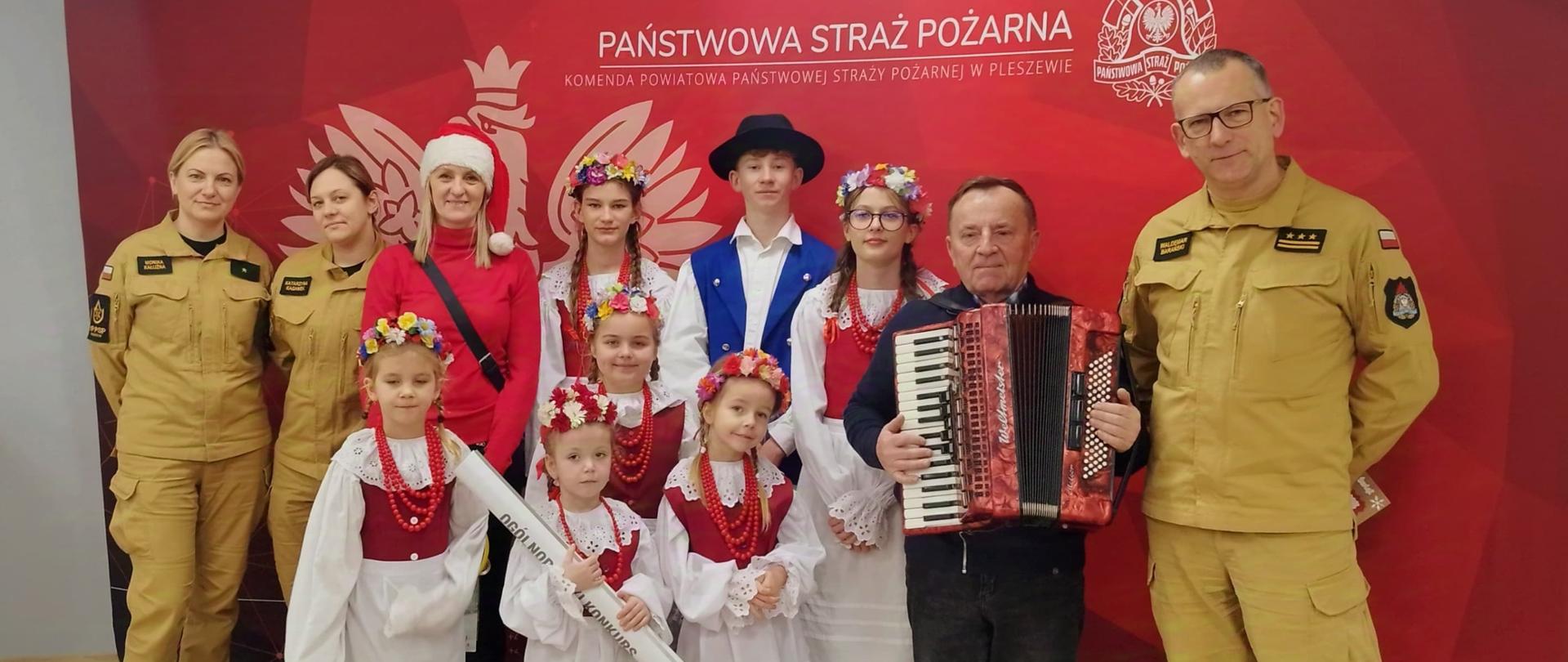 Na oświetlonej sali stoją dzieci ubrane w stroje ludowe, obok nich z prawej strony stoi mężczyzna w stroju ludowym z akordeonem oraz mężczyzna w żółtym mundurze Państwowej Straży Pożarnej, Z lewej strony stoją trzy kobiety, jedna w czerownym ubraniu cywilnym, dwie w żółtych mundurach Państwowej Straży Pożarnej, Za nimi widoczna jest czerwona ścianka reklamowa z logiem i napisem Państwowa Straż Pożarna