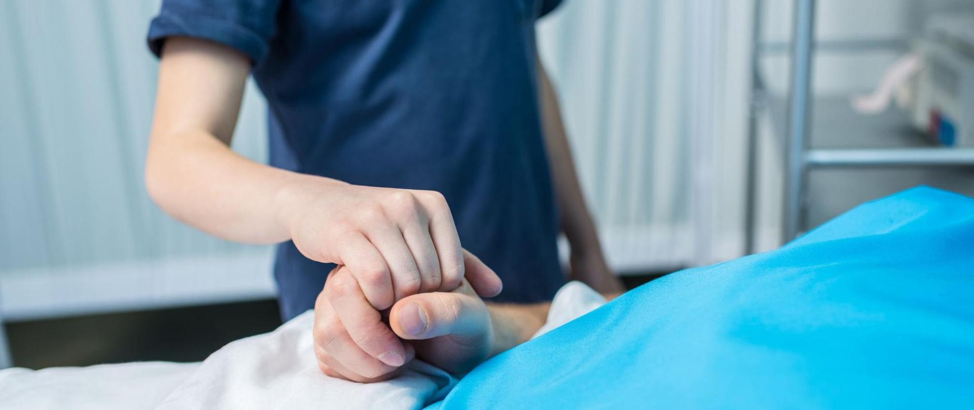Na zdjęciu jest leżący na łóżku pacjent oraz stojący nad nim lekarz trzymający go za rękę.
