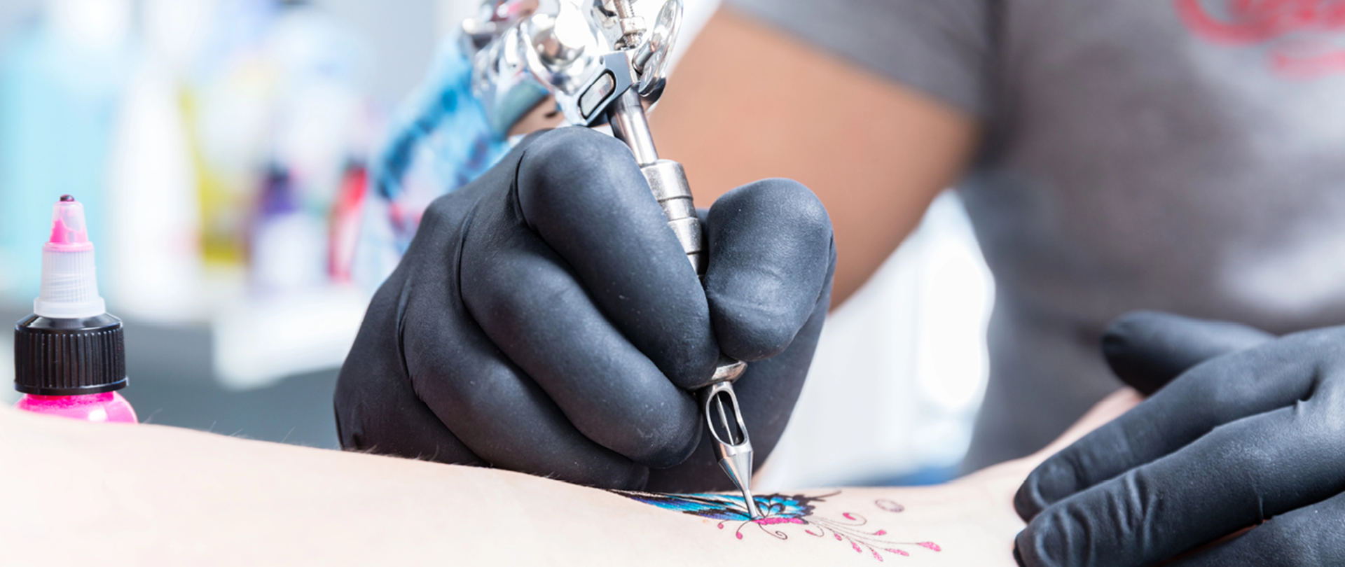 dwie ręce tatuażysty w czarnych lateksowych rękawiczka. W lewej ręce trzyma maszynkę do tatuażu. Pod ręką tatuażysty część ciała tatuowanej osoby, w tle buteleczka z farbą w kolorze różowym
