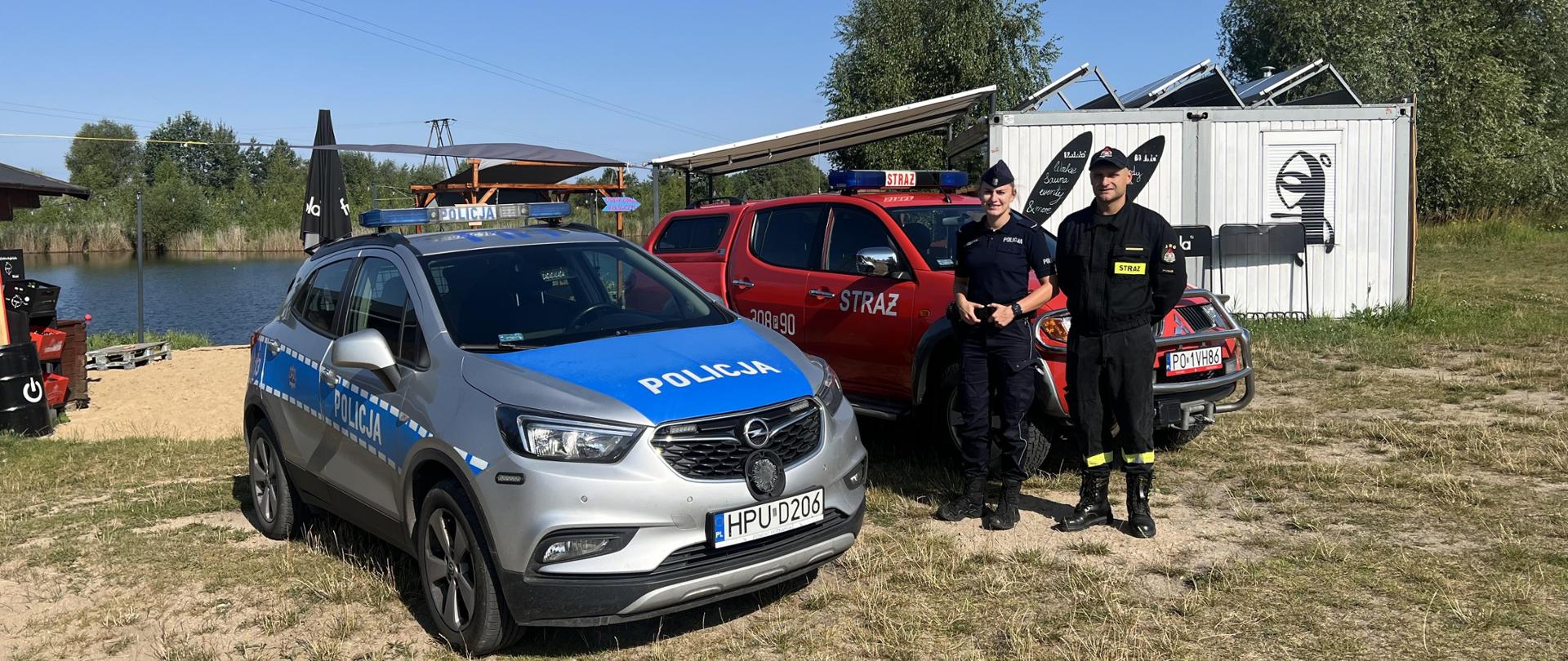 Samochód policyjny oraz strażacki. Obok samochodów stoją funkcjonariusze straży i policji, w tle widać akwen wodny Tropicana