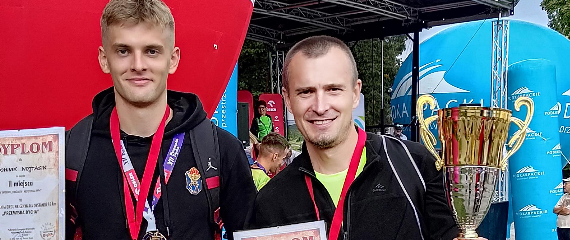 Zdjęcie pamiątkowe uczestników biegu. Dwóch mężczyzn z pamiątkowymi medalami na szyi, dyplomami w ręku (jeden z nich trzyma puchar) na tle czerwonego balonu z logo sponsora.