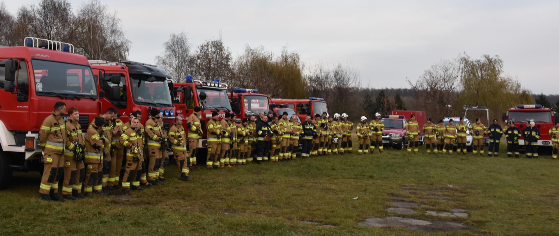 strażacy w trakcie zbiórki przed ćwiczeniami, w tle samochody pożarnicze