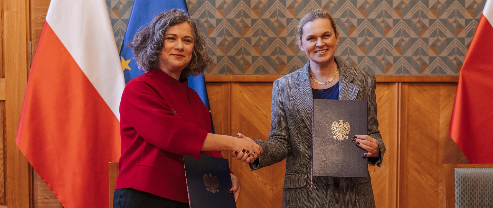 Duża sala ze ścianami w zielony wzorek na tle flag Polski i UE minister Nowacka podaje dłoń kobiecie z kobietą w czerwonej marynarce, obie panie w ręku trzymają granatowe teczki z grawerem orła. 