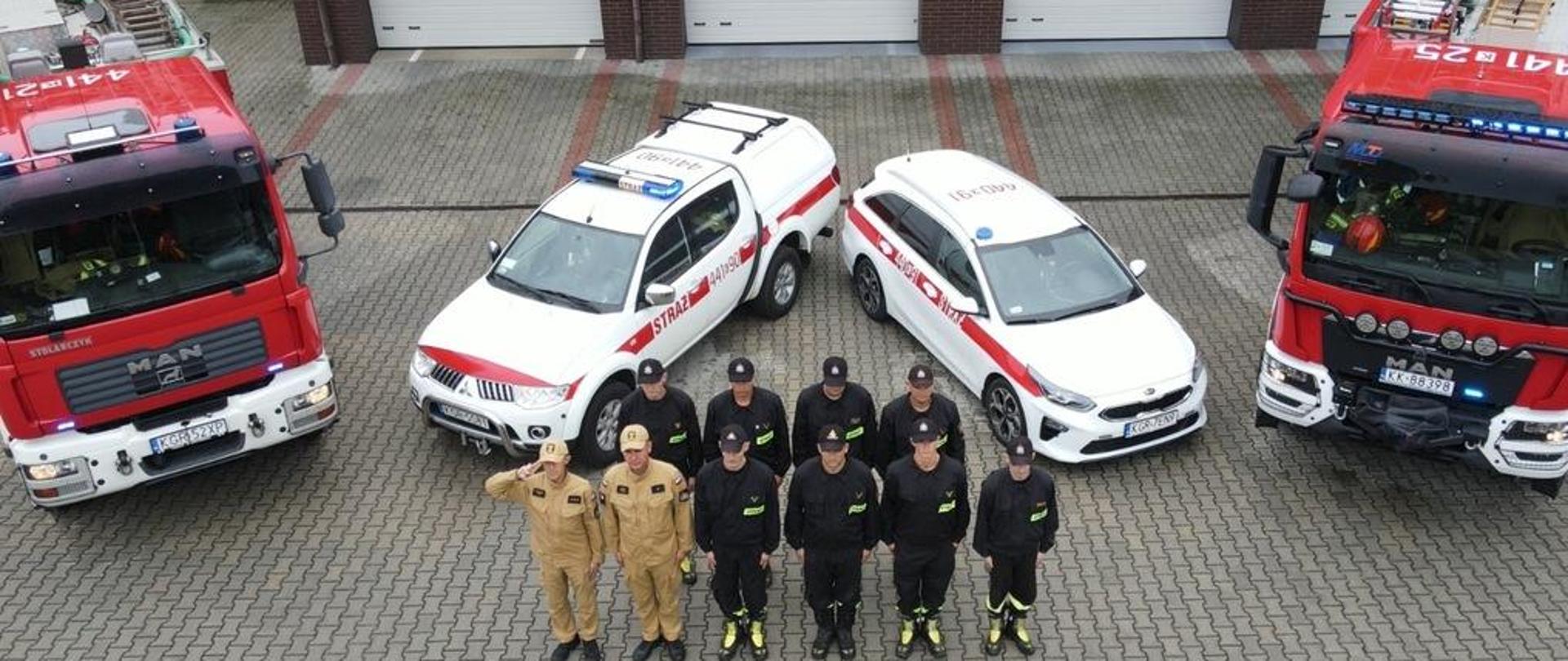 Na zdjęciu widoczni strażacy na tle budynku Komendy Powiatowej Państwowej Straży Pożarnej w Gorlicach za nimi samochody pożarnicze. Salutujący strażak dowódca zmiany służbowej po jego prawej stronie zmiana służbowa w dwuszeregu. Strażacy ubrani w ubrania koszarowe czarne w czapkach. Dowódca zmiany i strażak stojący obok w piaskowych ubraniach służbowych również w czapkach.