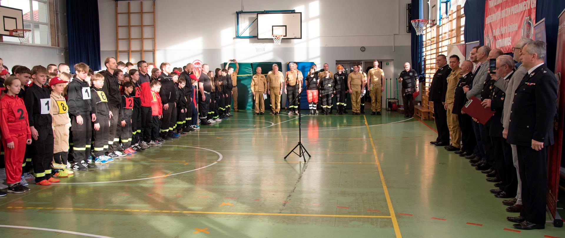 Ceremonia otwarcia turnieju na sali sportowej. Po lewej - drużyny, na środku - komisja sędziowska, po prawej - goście