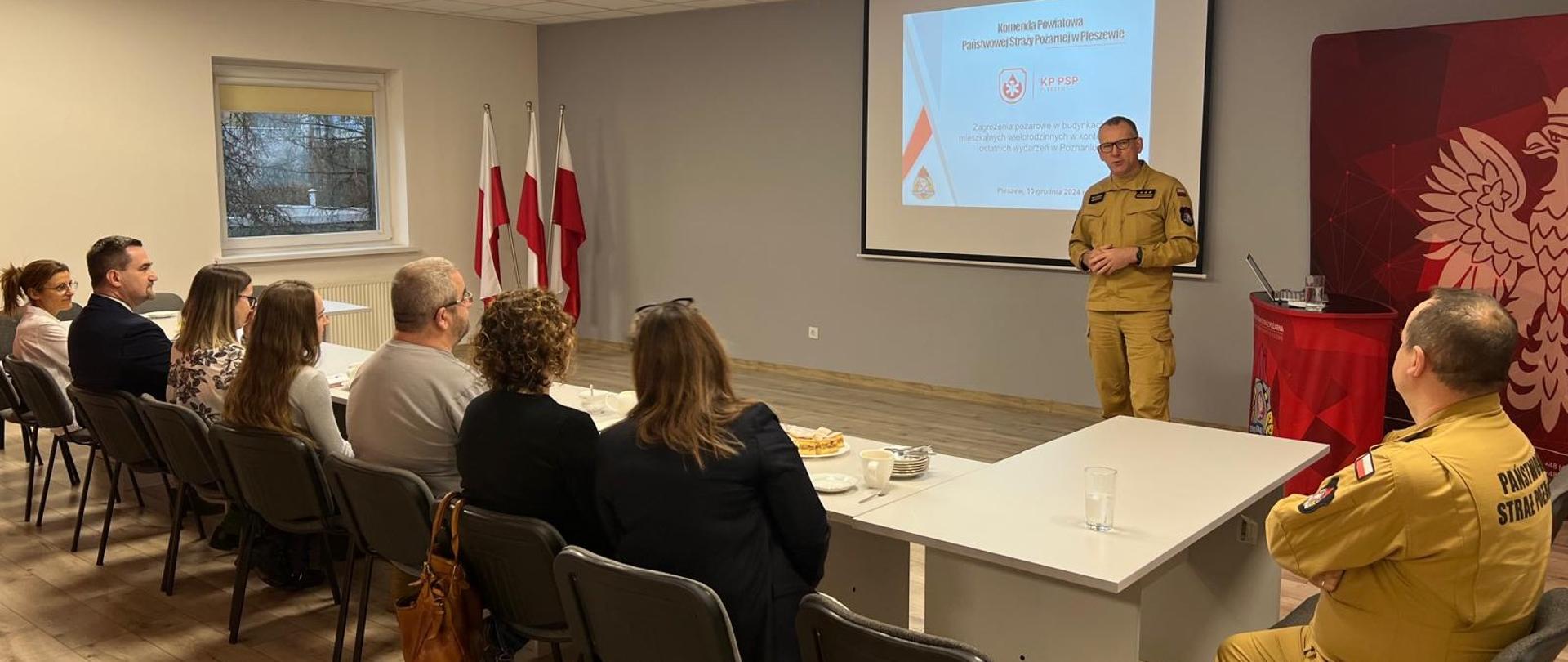 Na oświetlonej sali z ekranem, za stołami siędzą osoby w ubraniach cywilnych oraz mężczyzna w żółtym mundurze Państwowej Straży Pożarnej, przed nimi przemawia mężczyzna w żółtym mundurze Państwowej Straży Pożarnej, za nim widoczny jest czerwony baner z logiem i napisem Państwowa Straż Pożarna.