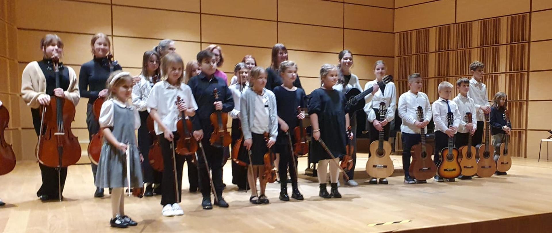 Grupa uczniów oraz nauczycieli ustawiona w dwóch rzędach na scenie sali koncertowej. Po prawej stronie uczniowie trzymają gitary, po lewej wiolonczele, a w środku skrzypce. 