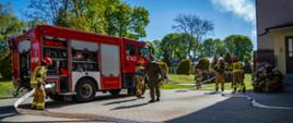 Zdjęcie zrobione w dzień. Na zdjęciu widać samochód strażacki, strażaków rozkładających sprzęt i żołnierzy stojących obok budynku. 