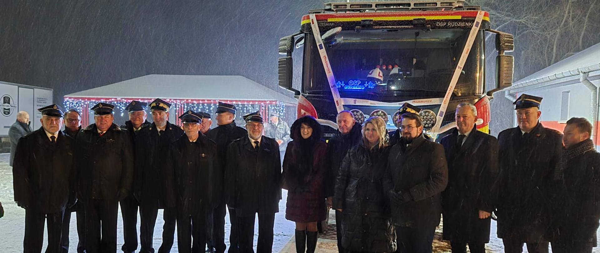 Zdjęcie grupy osób na tle przodu samochodu pożarniczego. W tle widoczne ośnieżone dachy budynków. Zdjęcie wykonane na przestrzeni otwartej w porze nocnej w czasie opadu śniegu. Plac apelowy oświetlony światłem sztucznym, które rzuca podłużne światłocienie.