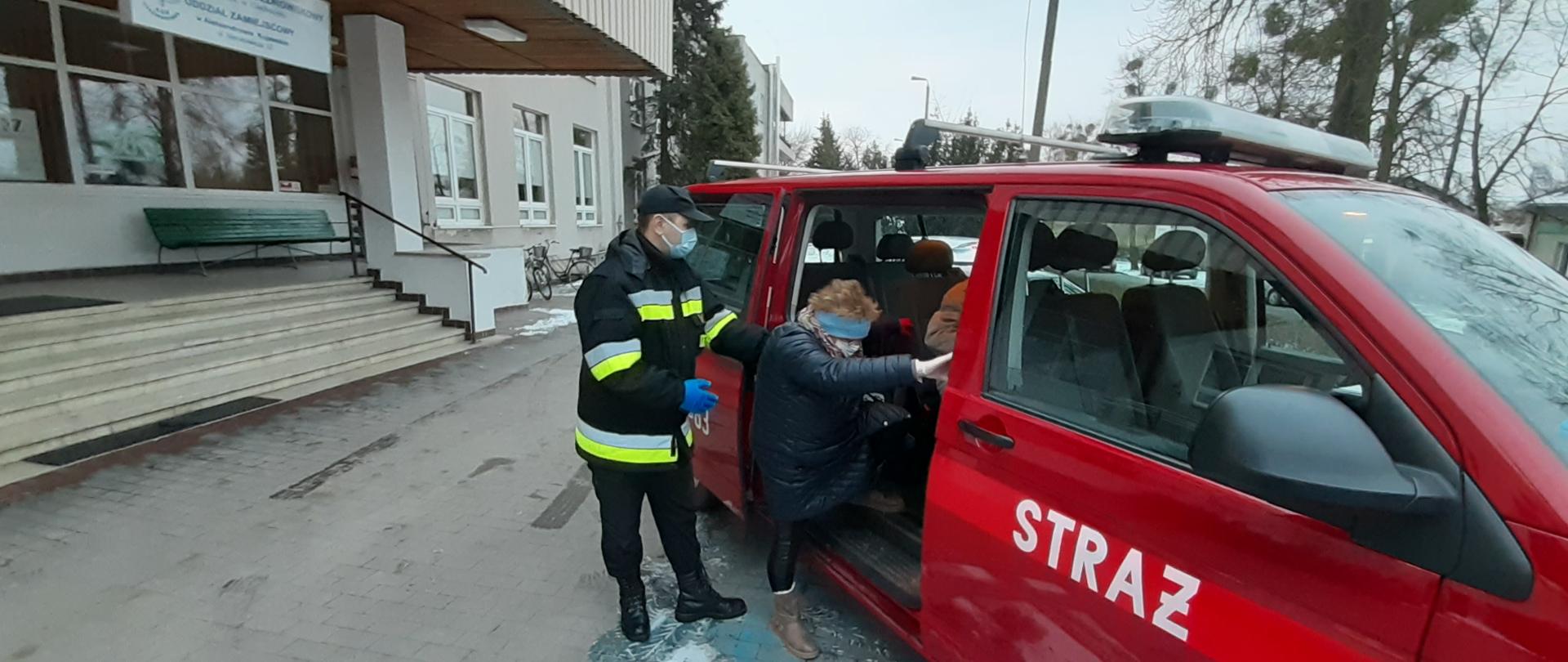 Na zdjęciu strażak pomaga wsiąść starszej kobiecie do strażackiego busa.