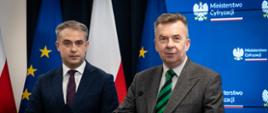 Na tle niebieskiej ścianki z napisami Ministerstwo Cyfryzacji stoi minister Wieczorek, obok mężczyzna w garniturze trzyma czarną teczkę z literami MC.