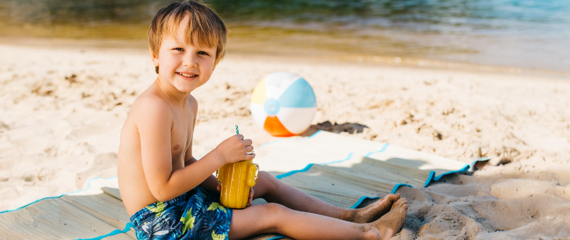 uśmiechnięty chłopiec siedzi na macie na plaży, w ręku trzyma kubek, obok piłka dmuchana