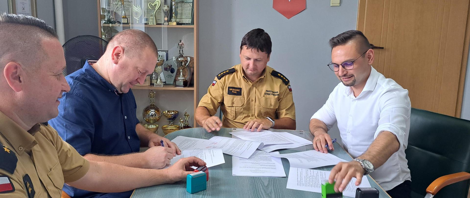 Zdjęcie przedstawia dwóch funkcjonariuszy PSP oraz samorządowców podpisujących dokumenty