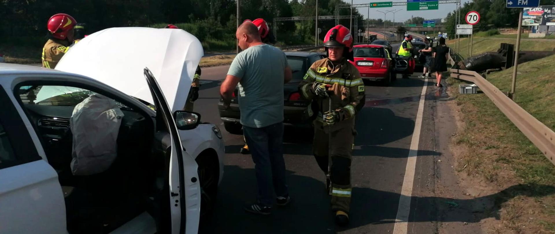 Zdjęcie przedstawia cztery samochody uczestniczące w kolizji stojące na poboczu jezdni. Obok samochodów widać osoby nieuczestniczące w kolizji oraz strażaków wykonujących czynności służbowe.