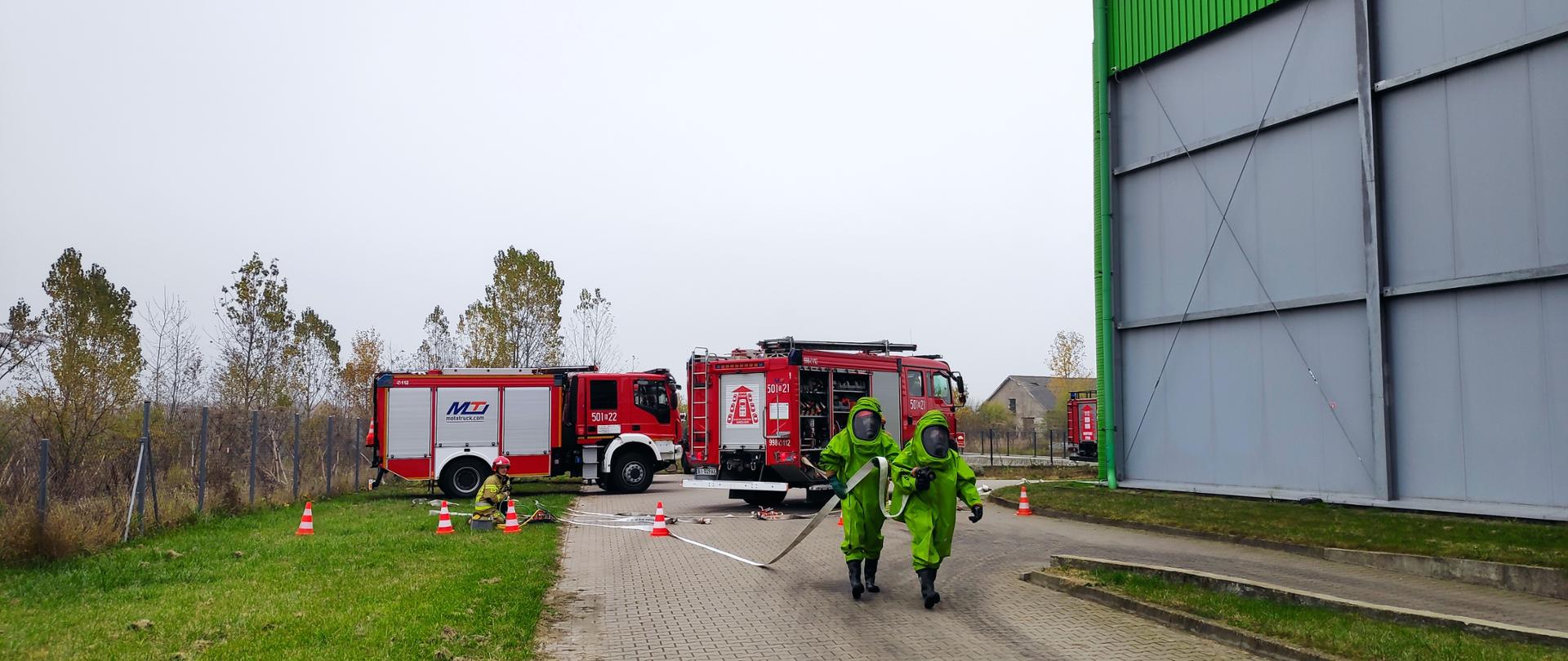 Zdjęcie przedstawia dwóch ratowników ubranych w ciężkie ubrania gazoszczelne, udających się do budynku w celu likwidacji zagrożenie. Z tyłu pracują strażacy ubrani w ubranie bojowe. Zdjęcie wykonano na terenie firmy BHJ w pochmurny dzień.