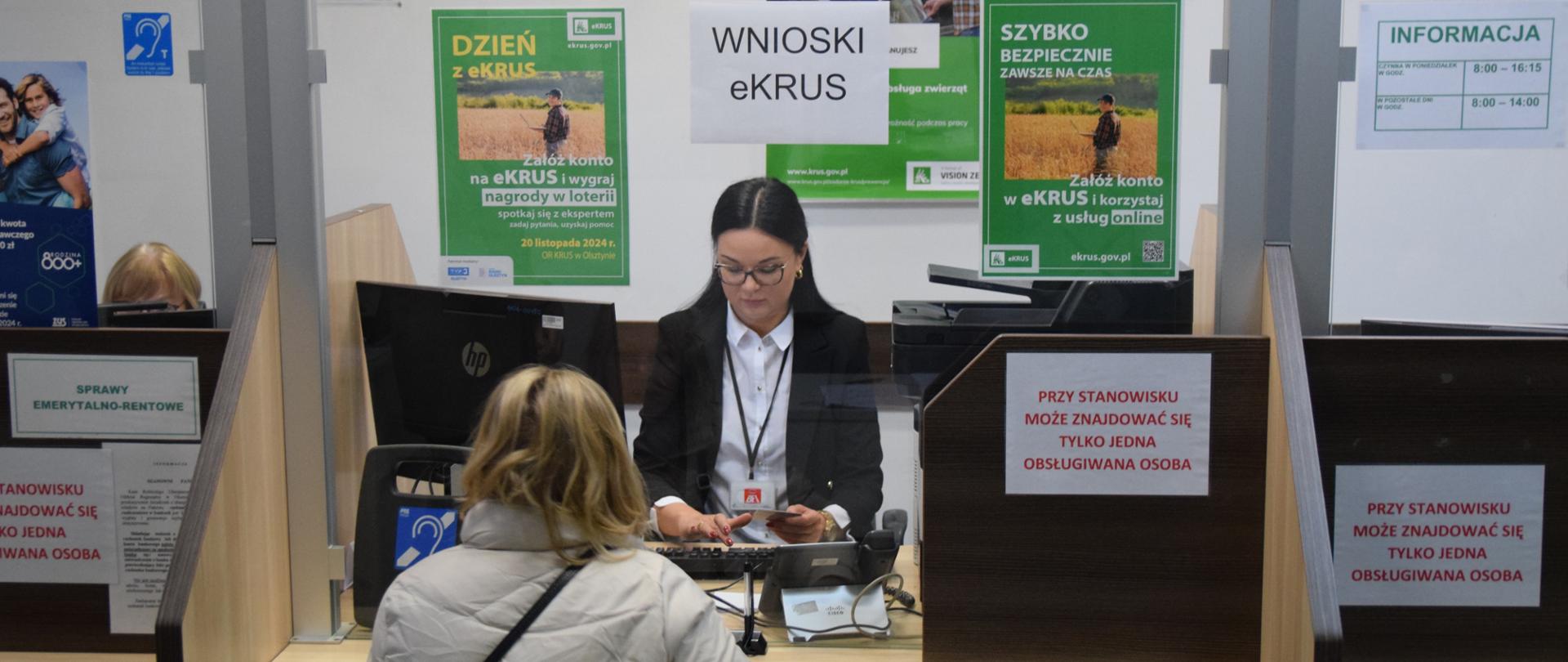 Kobieta obsługuje interesantkę w okienku KRUS. Z tyłu plakaty eKRUS.