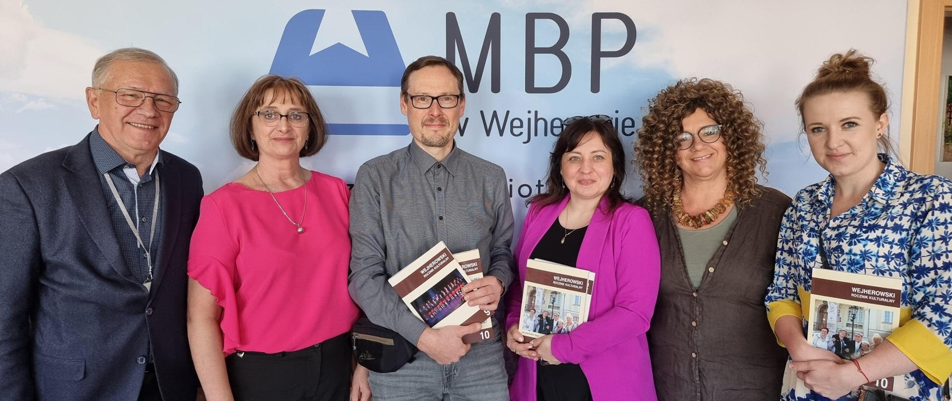 Na tle baneru Miejskiej Biblioteki Publicznej w Wejherowie pozują: redaktorzy rocznika, nauczyciele szkoły muzycznej oraz dyrektor Dorota Muża-Szlas.