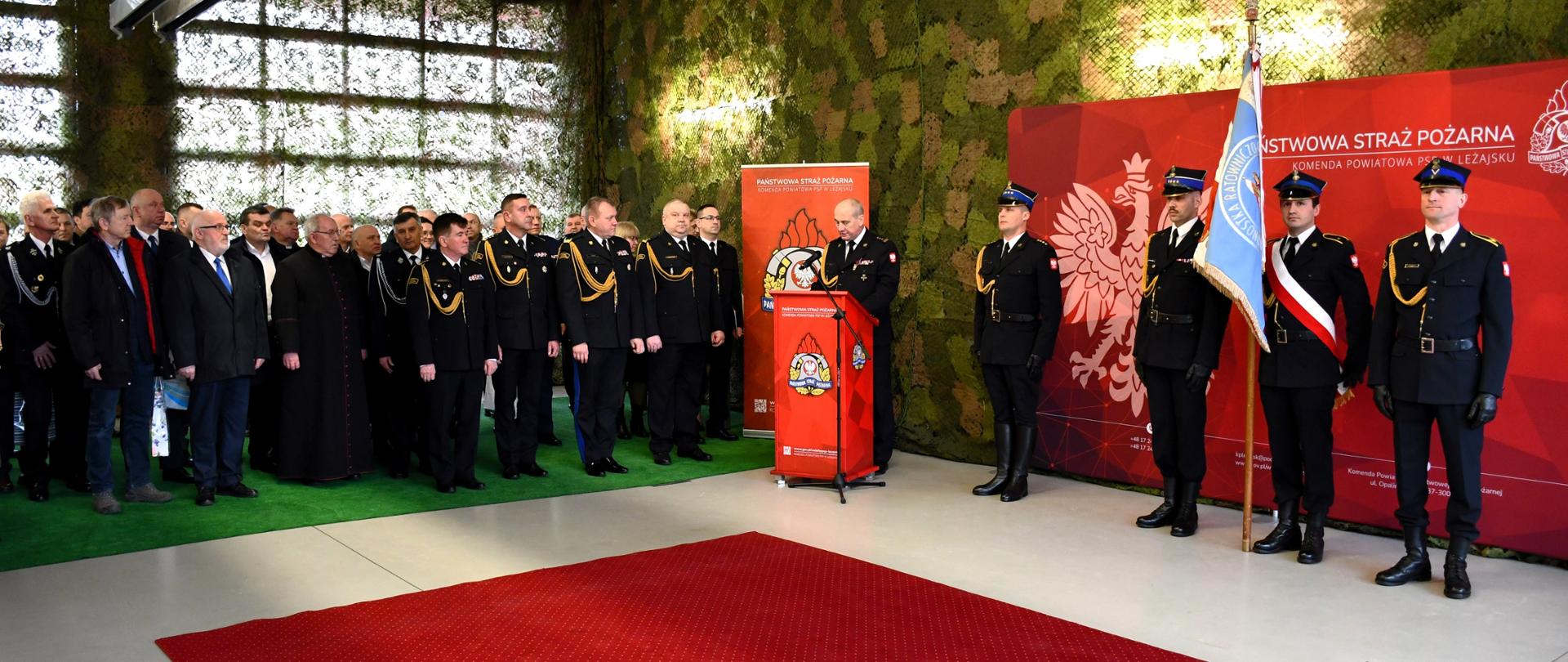 Na zdjęciu widzimy przemawiającego Komendanta Powiatowego PSP w Leżajsku. W tle widzimy zaproszonych gości oraz poczet sztandarowy.
