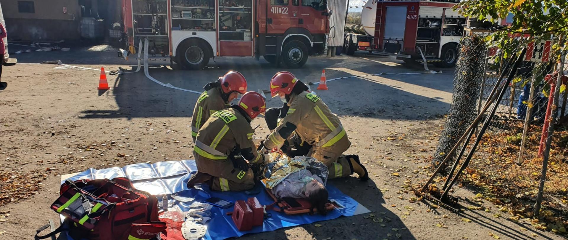 Zdjęcie przedstawia dwóch strażaków w umundurowaniu specjalnym, którzy udzielają pomocy medycznej osobie leżącej już na desce ortopedycznej. W tle znajdują się dwa samochody ratowniczo-gaśnicze. 