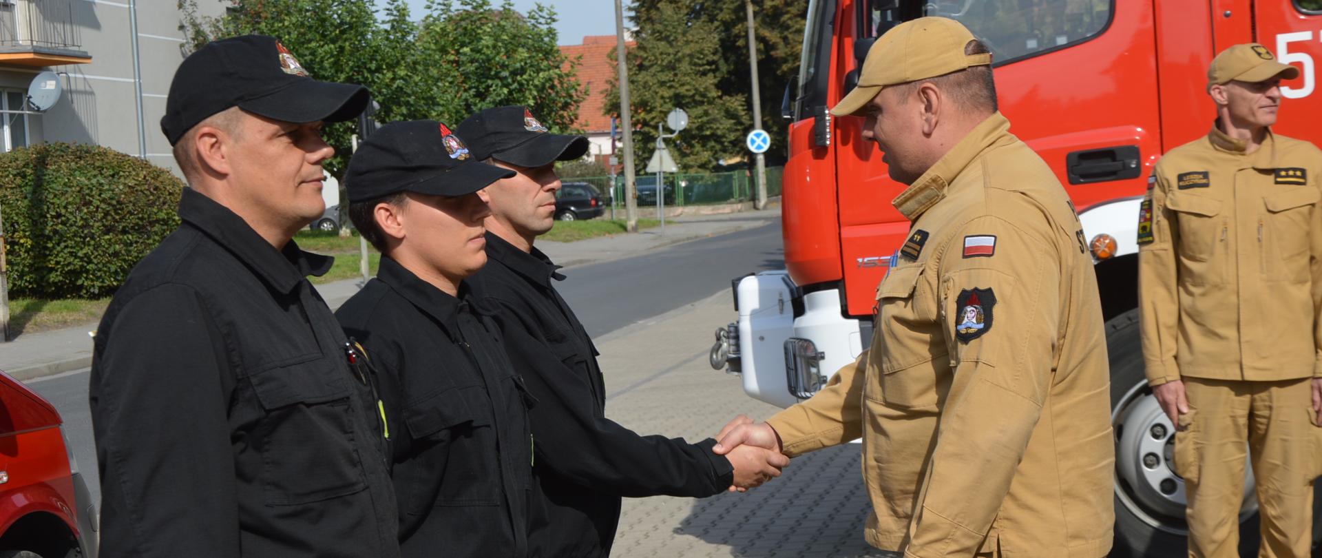 Plac manewrowy przed Komendą Powiatową PSP w Rawiczu. Na tle wozów pożarniczych komendant powiatowy poprzez uścisk dłoni składa podziękowanie i gratulacje trzem strażakom, którzy uczestniczyli w akcji przeciwpowodziowej. Świadkami tego wydarzenia są funkcjonariusze JRG. Strażacy mają na sobie ubrania koszarowe w kolorze ciemnym lub mundury służbowe w kolorze piaskowym. W tle budynki i drzewa.