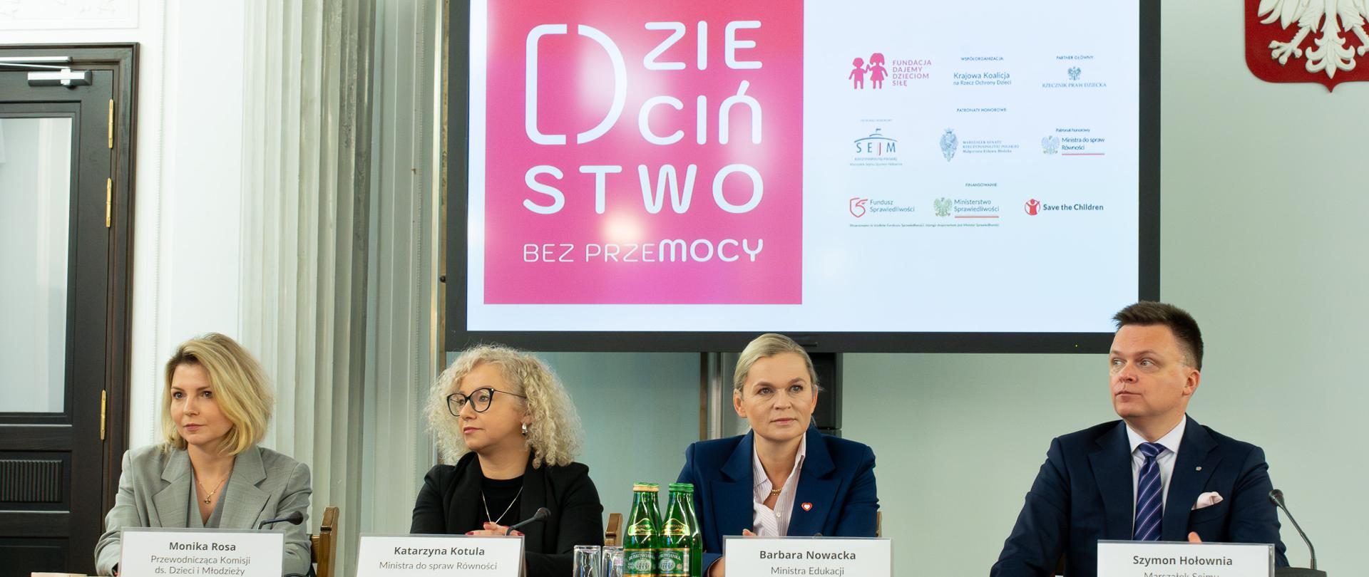 Przy stole siedzą od lewej: Monika Rosa, Katarzyna Kotula, Barbara Nowacka i Szymon Hołownia. Za nimi jest ustawiony telewizor na którym wyświetlany jest tekst: Dzieciństwo bez przemocy oraz logotypy partnerów biorących udział w akcji. 