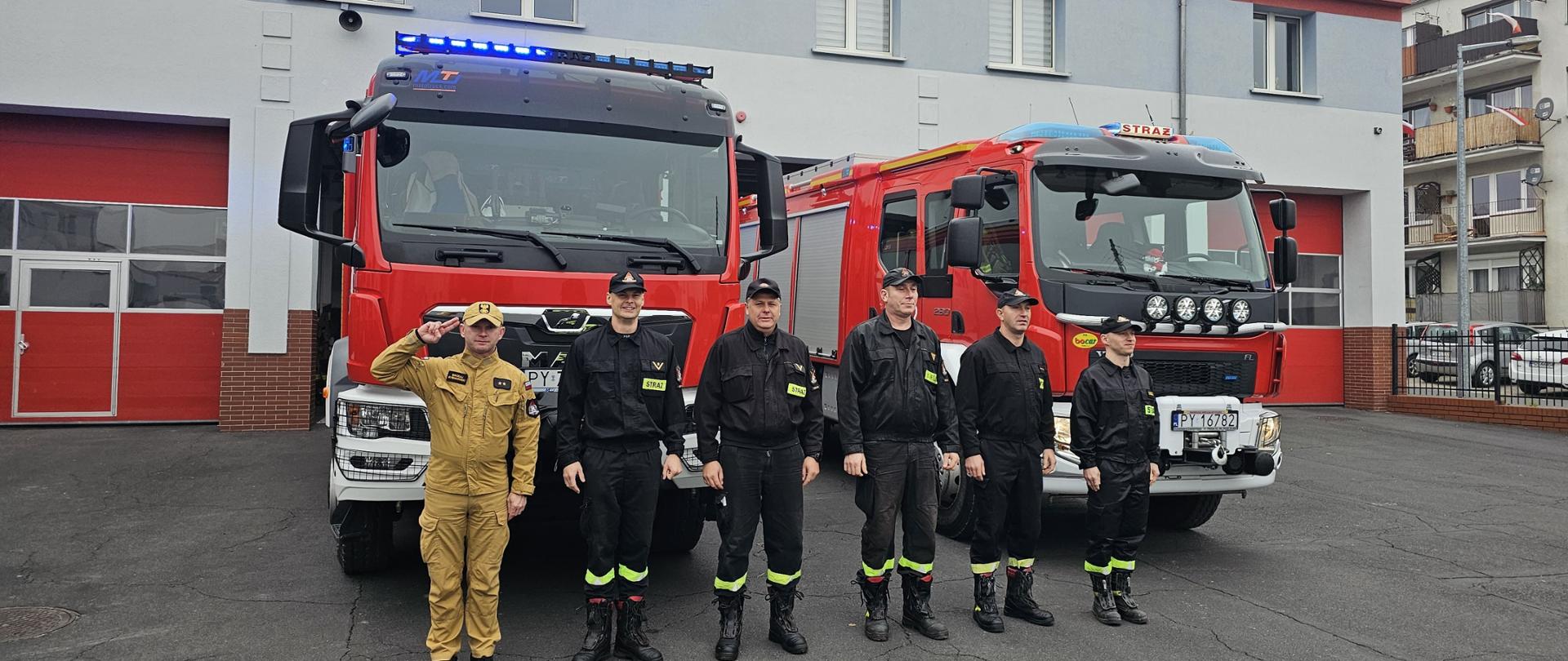 Strażacy stoją przed dwoma pojazdami pożarniczymi, przed komendą.