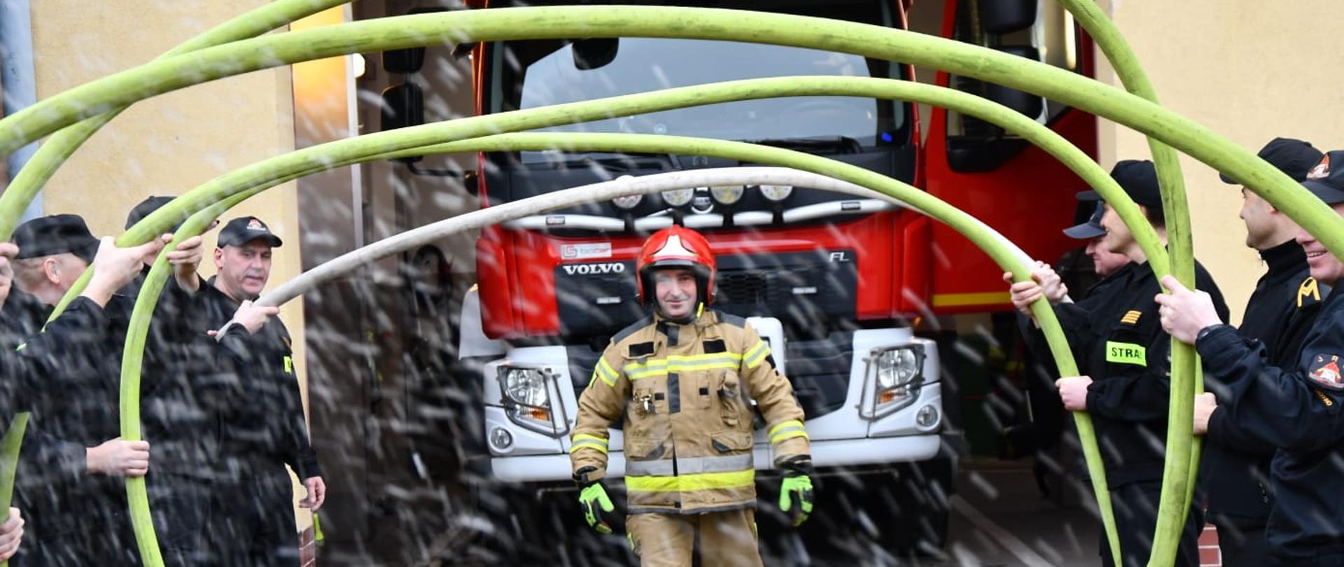 Zdjęcie przedstawia przechodzącego przez szpaler z węża pożarniczego starszego ogniomistrza w stanie spoczynku Krzysztofa Lutyńskiego