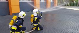 Strażacy z OSP Bucz podczas egzaminu praktycznego. Wyposażeni w aparaty powietrzne prezentują swoje umiejętności z zakresu gaszenia pożarów wewnętrznych. Jeden z nich, podaje prąd wody nad siebie.