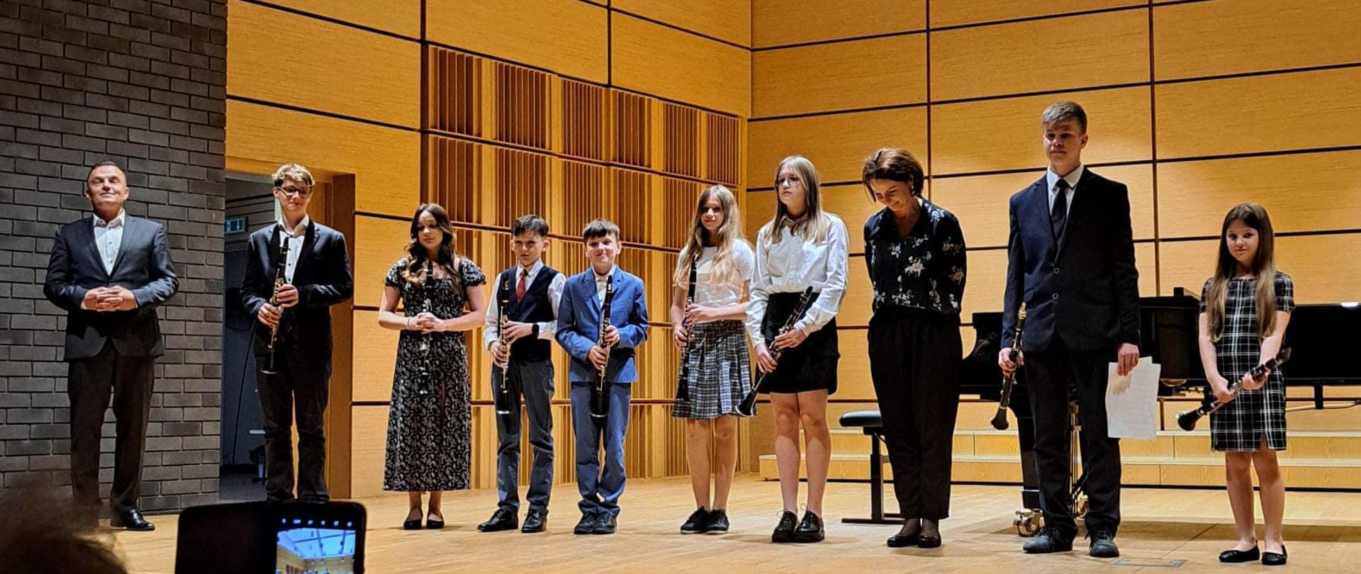 Uczniowie wraz z nauczycielami stojący w jednym rzędzie na scenie sali koncertowej. Każdy z uczniów trzyma klarnet. W tle, po prawej stronie widoczny jest fortepian. Z sufitu zwisają lampy.