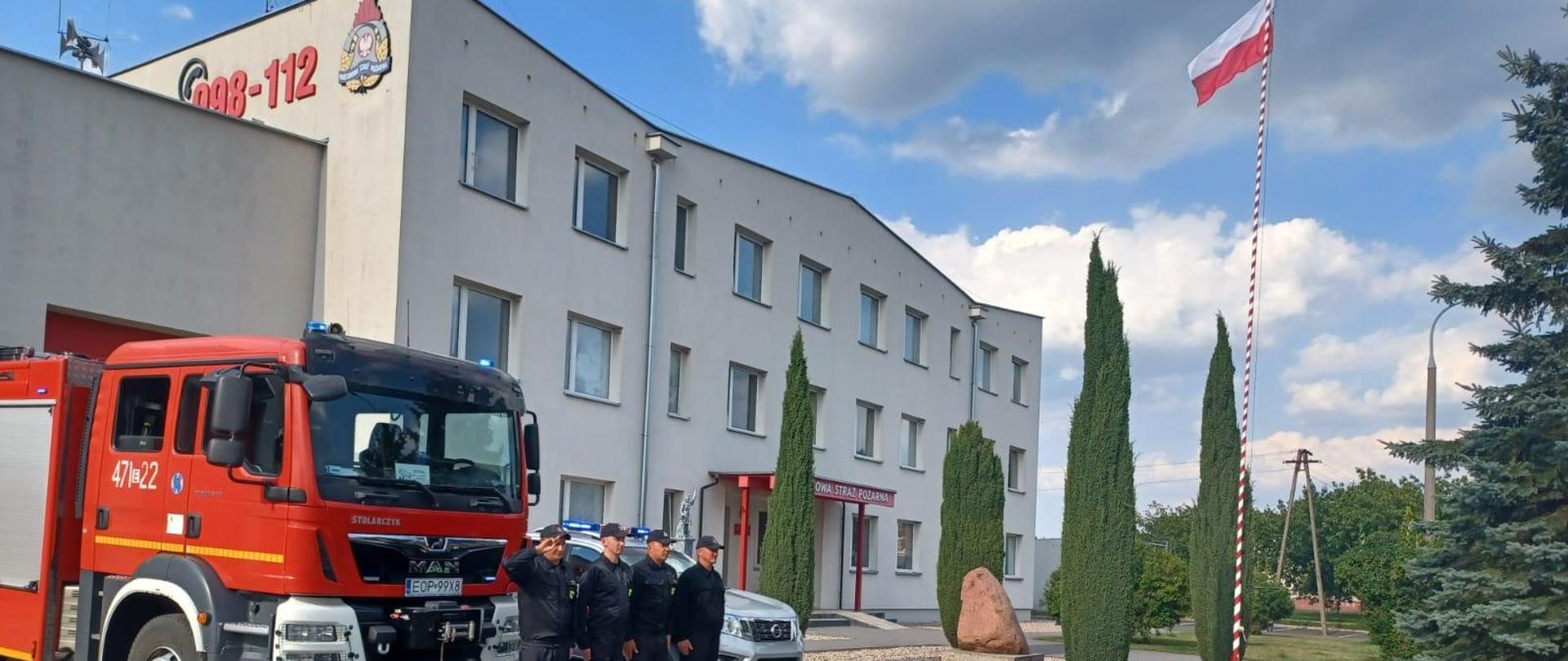 Czterech strażaków w umundurowaniu koszarowym na tle samochodów pożarniczych przed budynkiem komendy koloru szarego. Obok powiewająca biało - czerwona flaga na maszcie.