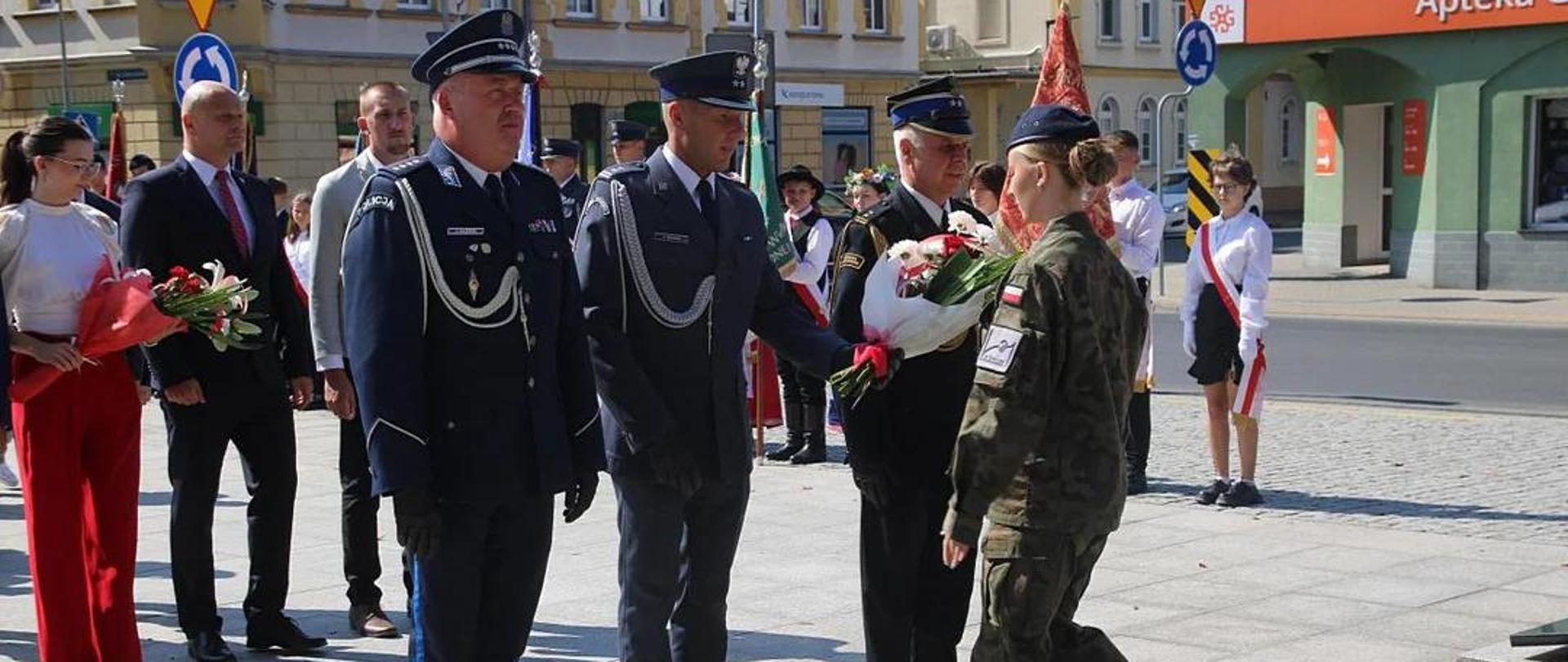 85. rocznica wybuchu II Wojny Światowej Plac przed pomnikiem Żołnierza Polskiego. Delegacja awickich służb mundurowych w składzie: policjant, funkcjonariusz Słuzby Więziennej i strażak, składa wiązankę kwiatów. Kwiaty uczennicy klasy mundurowej przekazuje funkcjonariusz SW. W tle pozostałe delegacje, poczty sztandarowe oraz zabudowania.