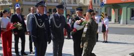 Plac przed pomnikiem Żołnierza Polskiego. Delegacja awickich służb mundurowych w składzie: policjant, funkcjonariusz Słuzby Więziennej i strażak, składa wiązankę kwiatów. Kwiaty uczennicy klasy mundurowej przekazuje funkcjonariusz SW. W tle pozostałe delegacje, poczty sztandarowe oraz zabudowania.