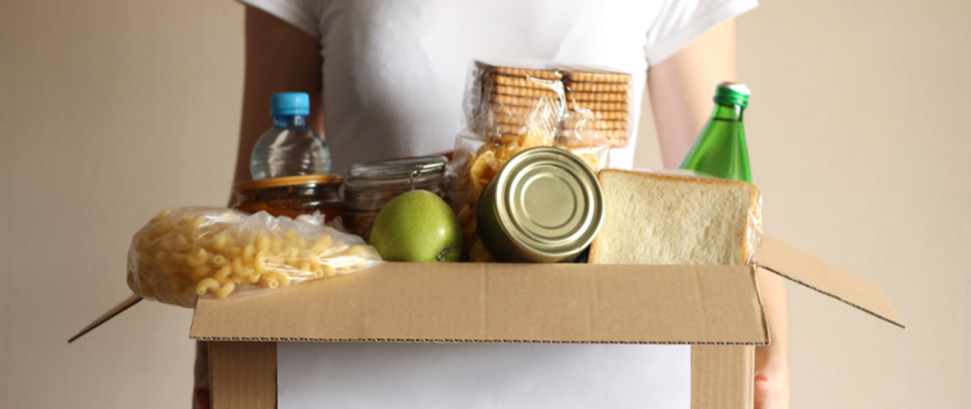 Na zdjęciu znajduje się kobieta, której twarzy nie widać trzymająca karton z różnymi produktami żywnościowymi 