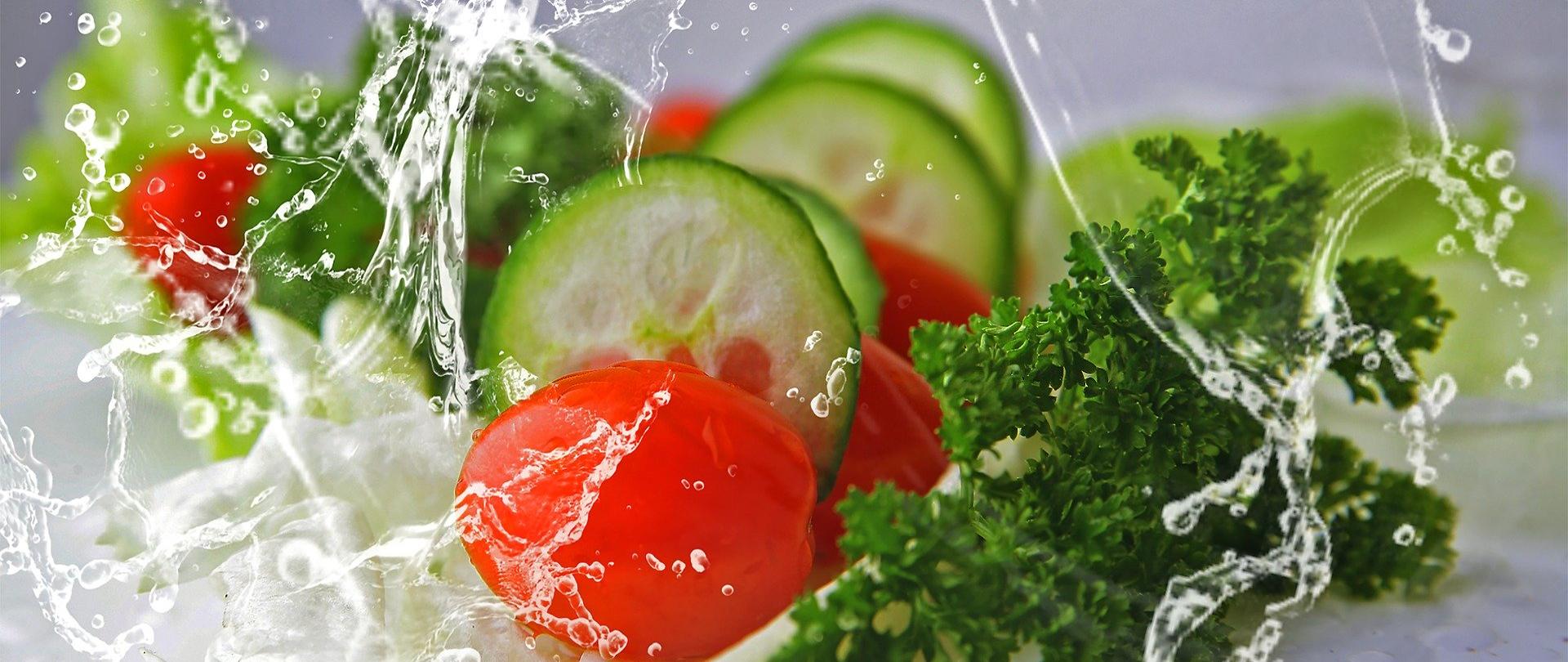 Na zdjęciu znajdują się warzywa tj. ogórek, pomidor i natka pietruszka oblane strumieniem wody. Tło jest szare.