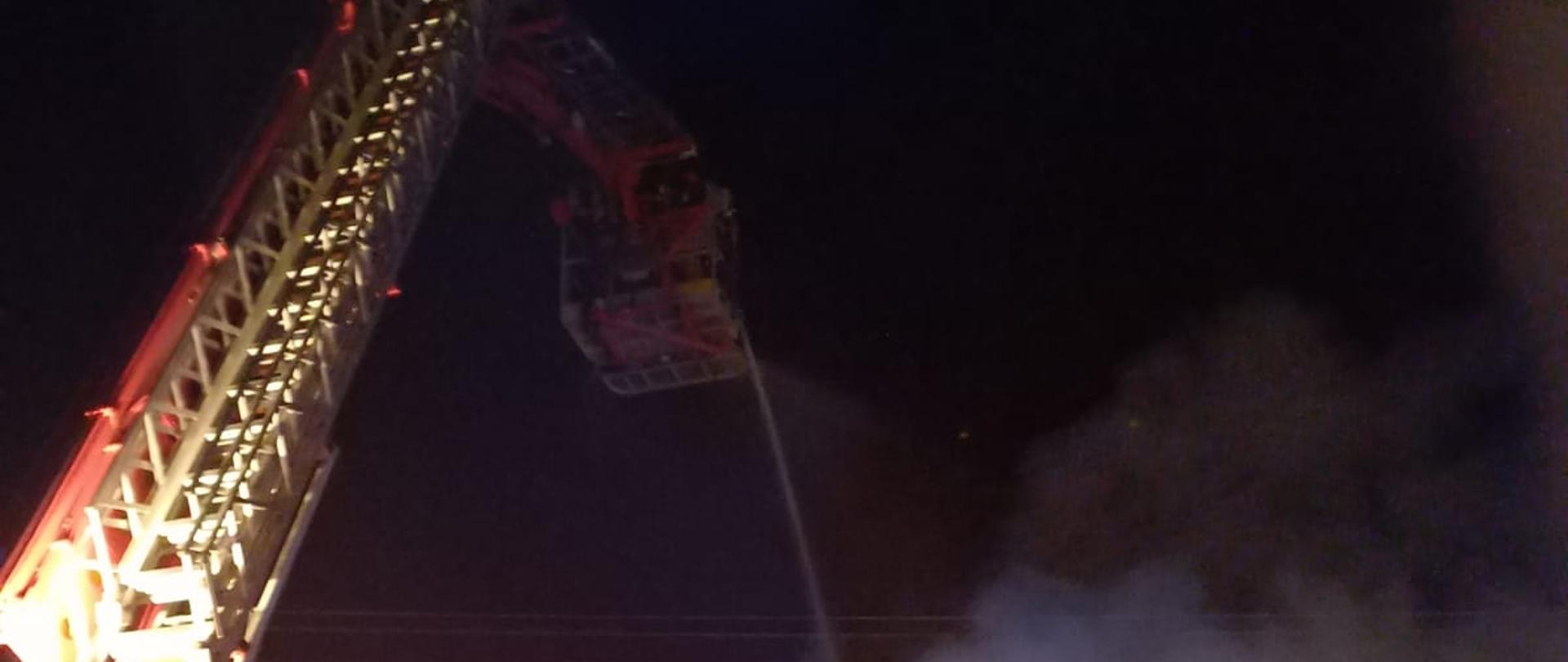 Po prawej stronie fotografii stojąca strażacy PSP, po lewej stronie widać tył podnośnika hydraulicznego, w tle palący się dom jednorodzinny, od góry strażacy PSP gaszą palący się dom z kosza podnośnika hydraulicznego. 