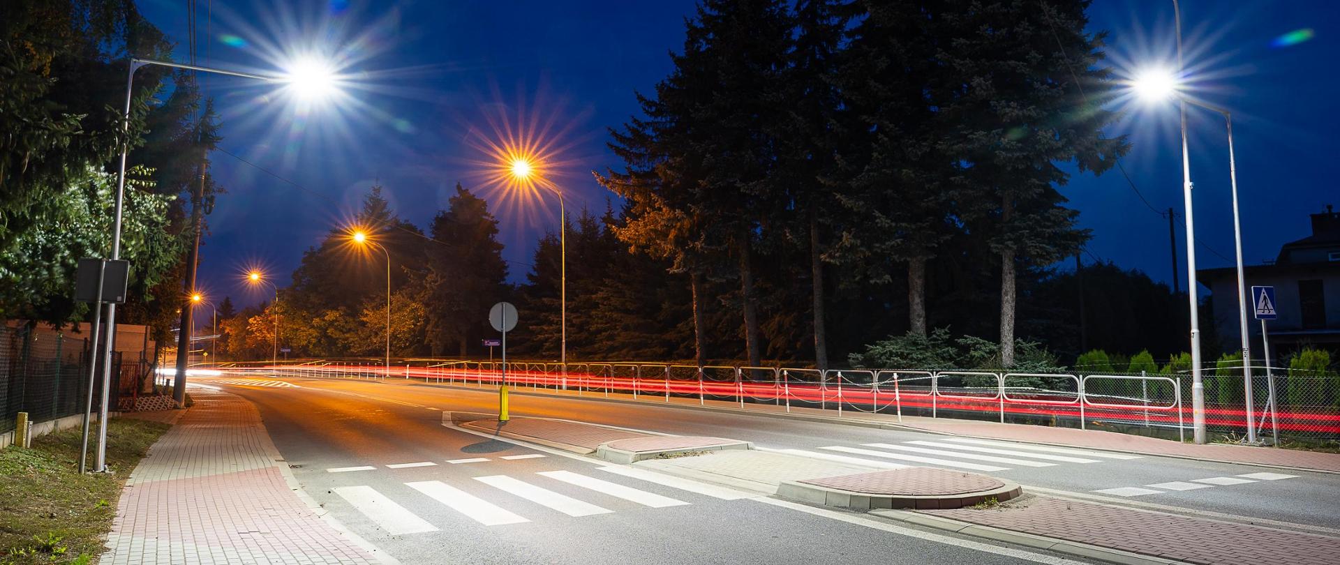 Widok na przejście dla pieszych w Boguchwale DK19. Pora nocna, przejście dla pieszych doświetlone, chodnik i przejście dla pieszych z wyspą rozdzielającą na środku.