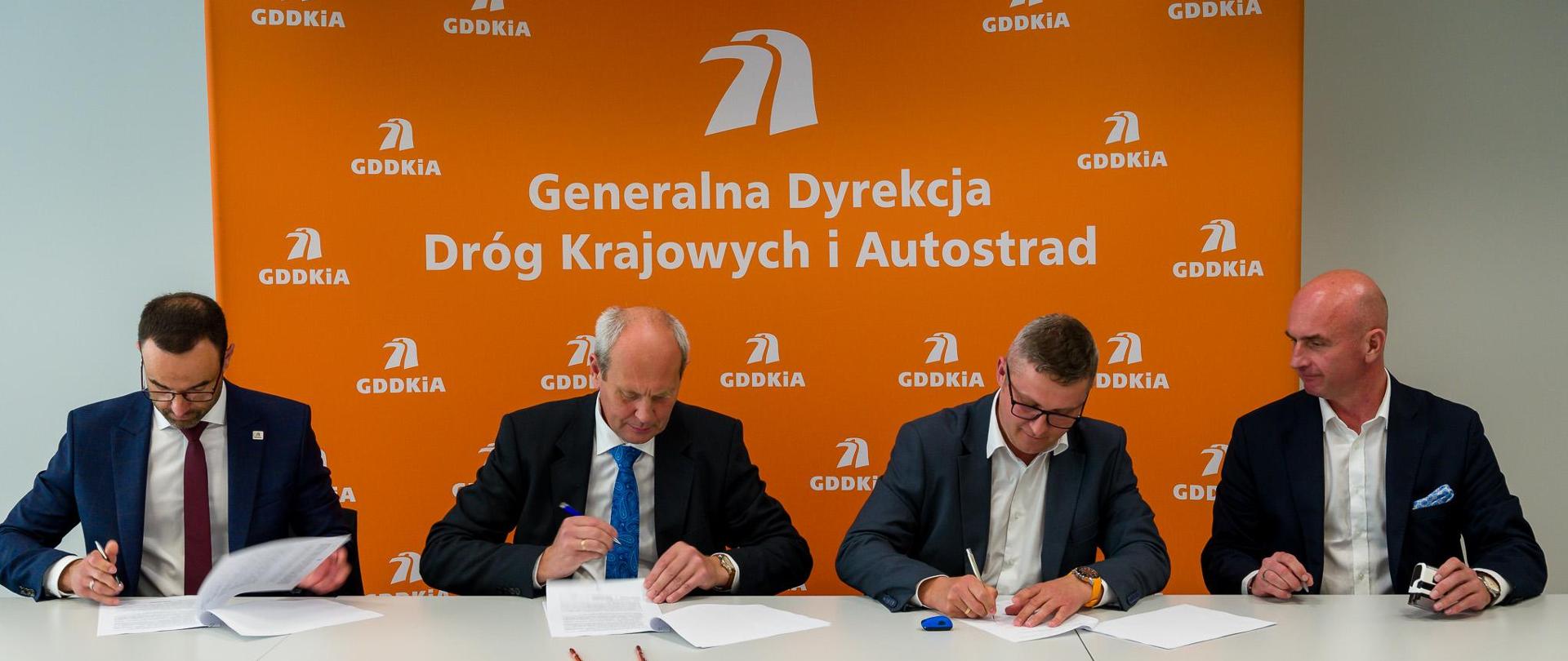 Moment podpisania jednego z 18 aneksów waloryzacyjnych. 4 elegancko ubranych mężczyzn siedzących obok siebie podpisuje dokumenty. W tle pomarańczowa ścianka z logotypami i nazwą GDDKiA.