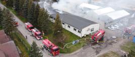 Zdjęcie przedstawia pożar hali produkcyjnej. Dookoła stoją wozy strażackie i prowadzone są działanie gaśnicze.
