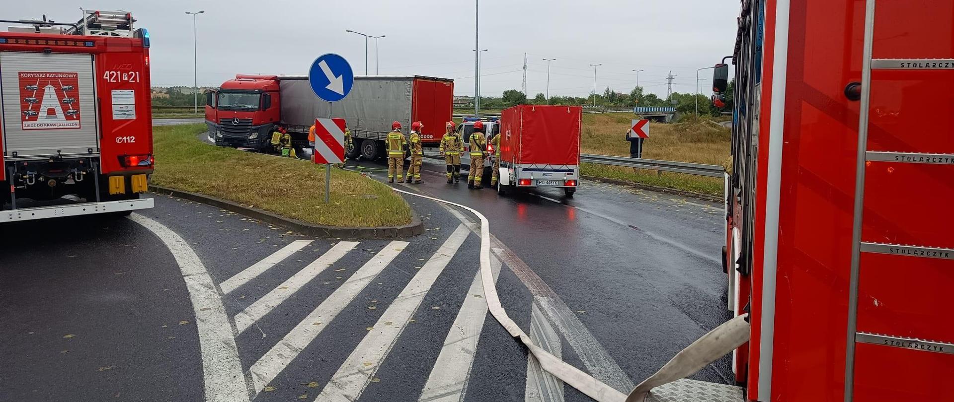 Na zdjęciu widać samochody pożarnicze oraz strażaków obsługujących zdarzenie. W tle widać samochód ciężarowy, który na łuku zjazdu „złamał się” blokując całkowicie przejazd. Samochód znajduje się w pozycji na kołach.