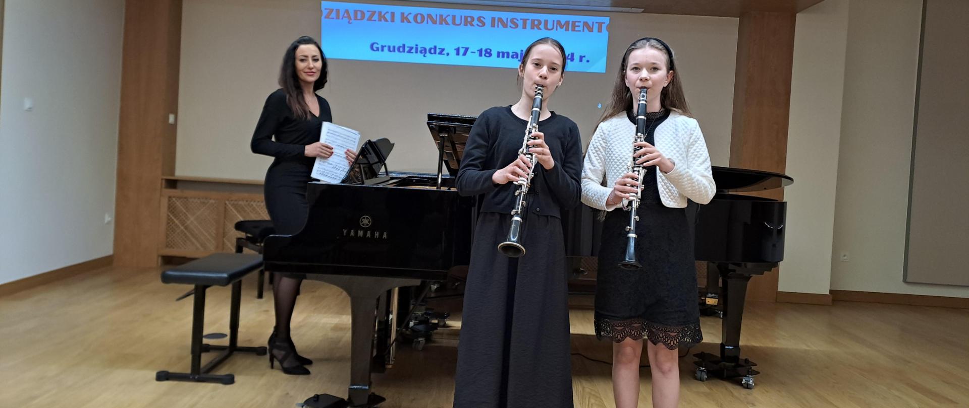 Na sali koncertowej w Grudziądzu pozują z klarnetami dwie uczennice. Za nimi znajduje się fortepian a przy nim stoi akompaniatorka.