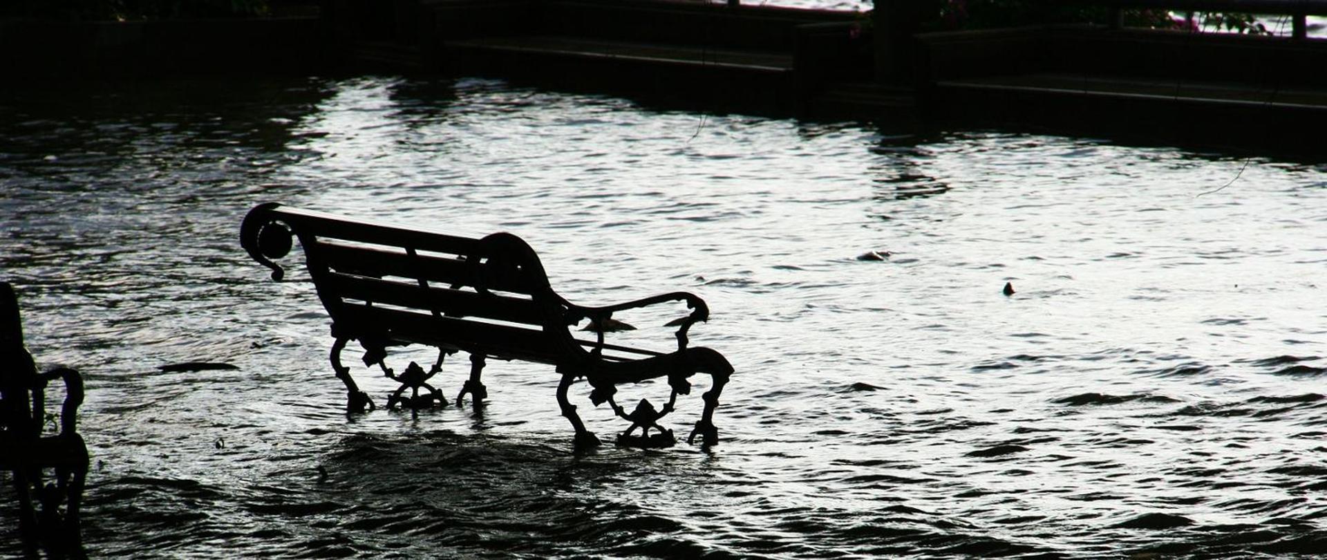 Na zdjęciu znajduje się ławka, która stoi w wodzie. Zdjęcie obrazuje zalanie spowodowane gwałtowną ulewą.