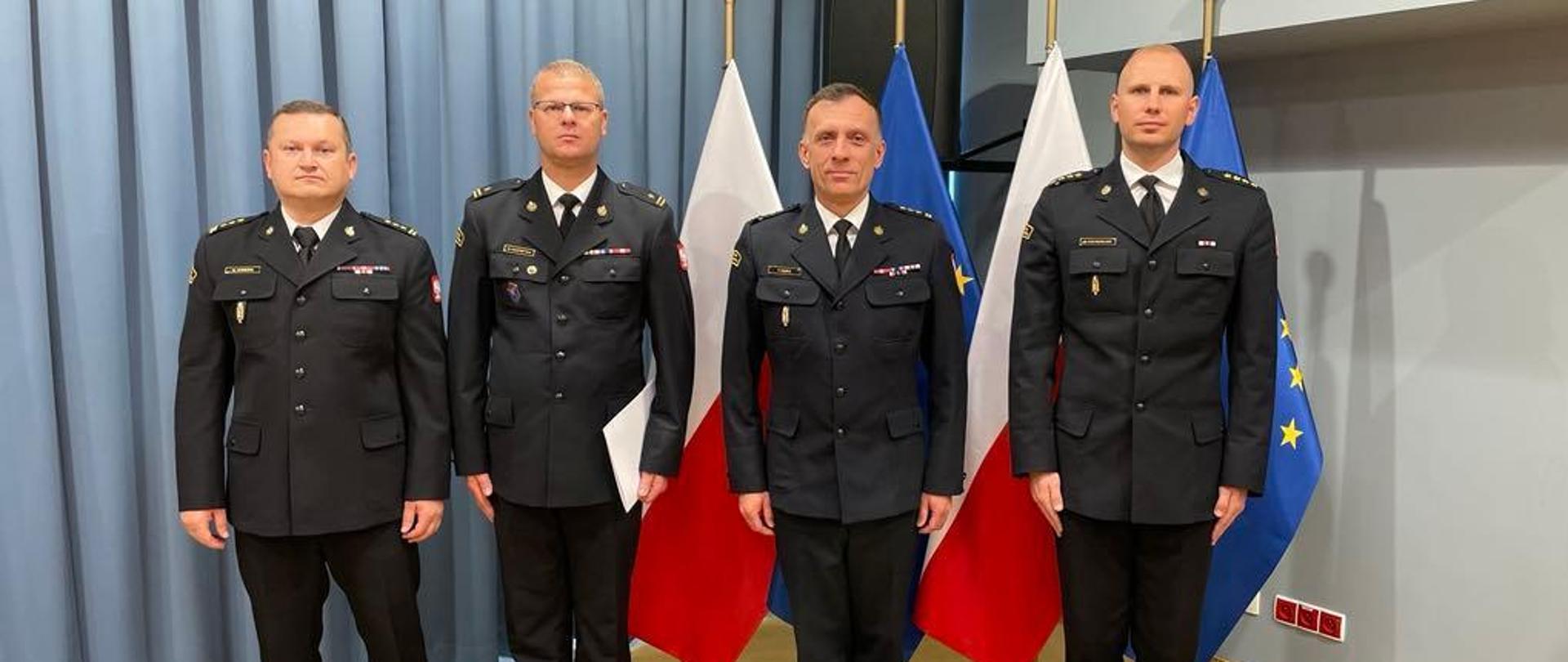 Czterech funkcjonariuszy w mundurach wyjściowych stoi w szeregu w pomieszczeniu, jeden z nich trzyma w ręku białą teczkę. Za nimi stoją flagi Polski i Uni Europejskiej. 