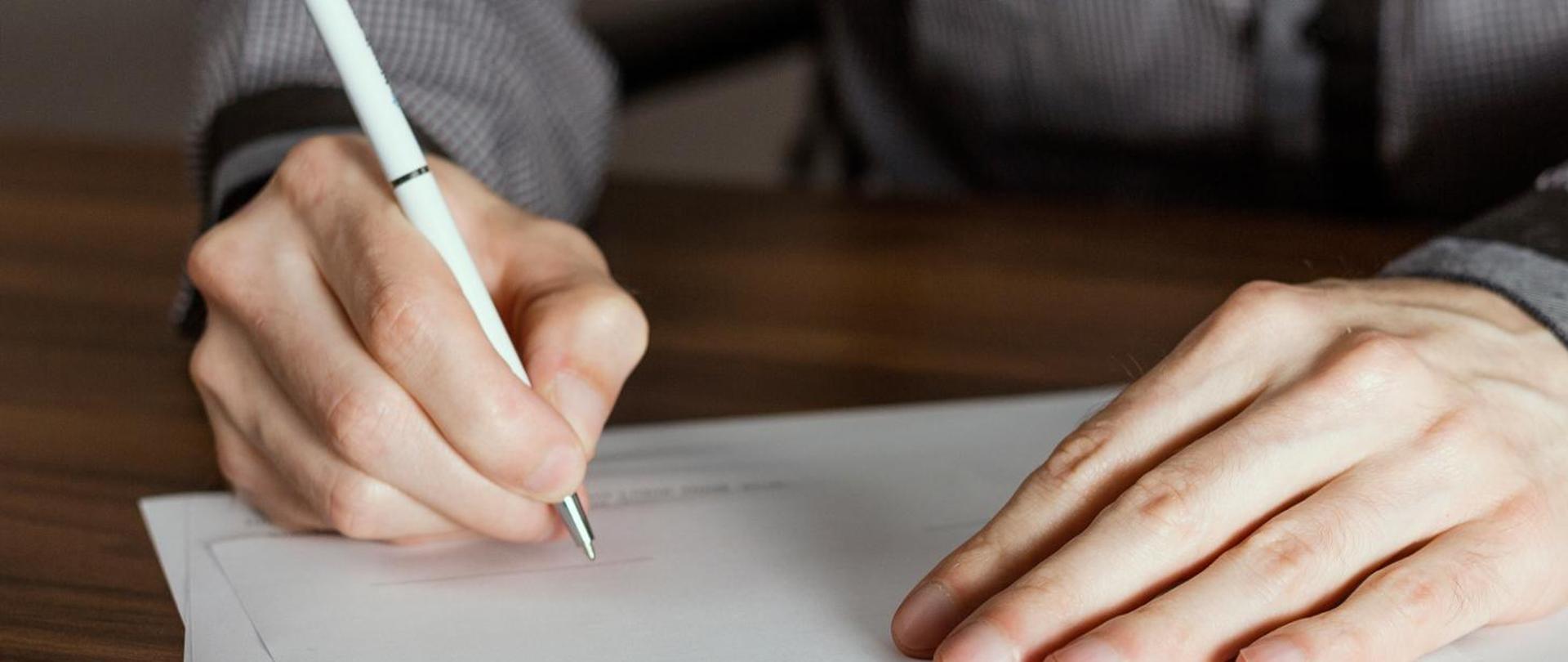 Na zdjęciu widać dłonie oparte o stolik. Jedna dłoń trzyma biały długopis i pisze po kartce papieru.