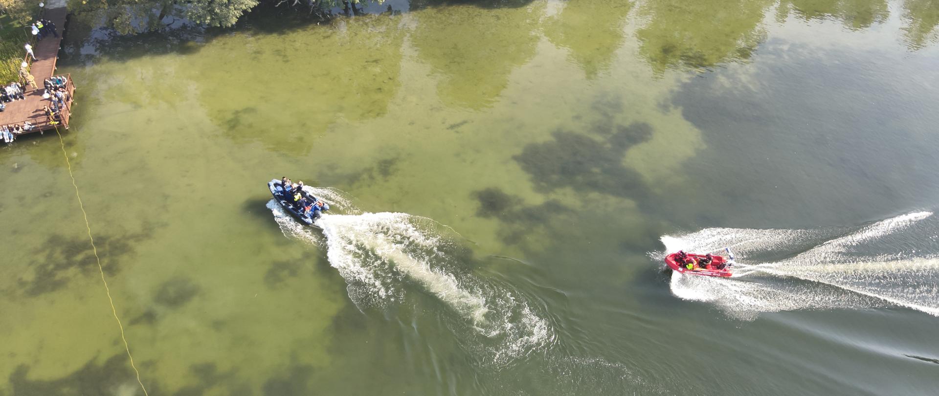 Fotografia przedstawia ujęcie wykonane z wysokości 30 metrów z drona. Widoczne dwie łodzie: policyjna niebieska i strażacka czerwona oraz dron podwodny połączony kabloliną z operatorem. Sprzęt znajduje się na wodzie niedaleko brzegu i pomostu, na którym stoją obserwatorzy – młodzież.