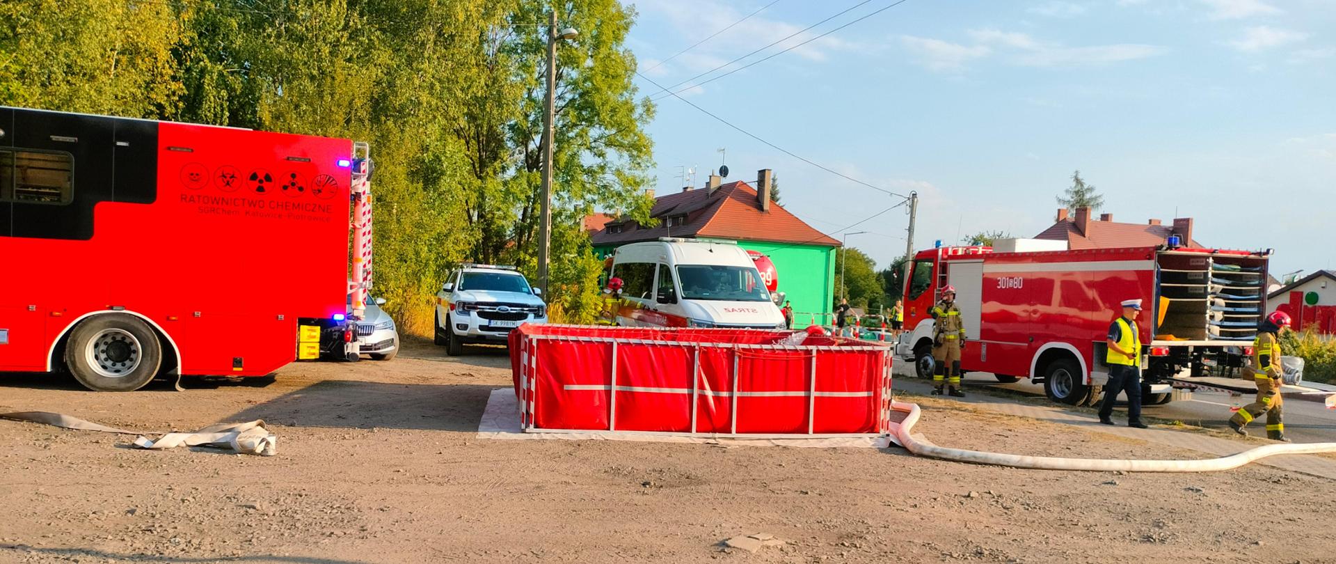 W tle wóz chemiczny Państwowej Straży Pożarnej, wóz pożarniczy oraz strażacy i funkcjonariusze policji, na pierwszym planie basen do którego wlewana jest woda