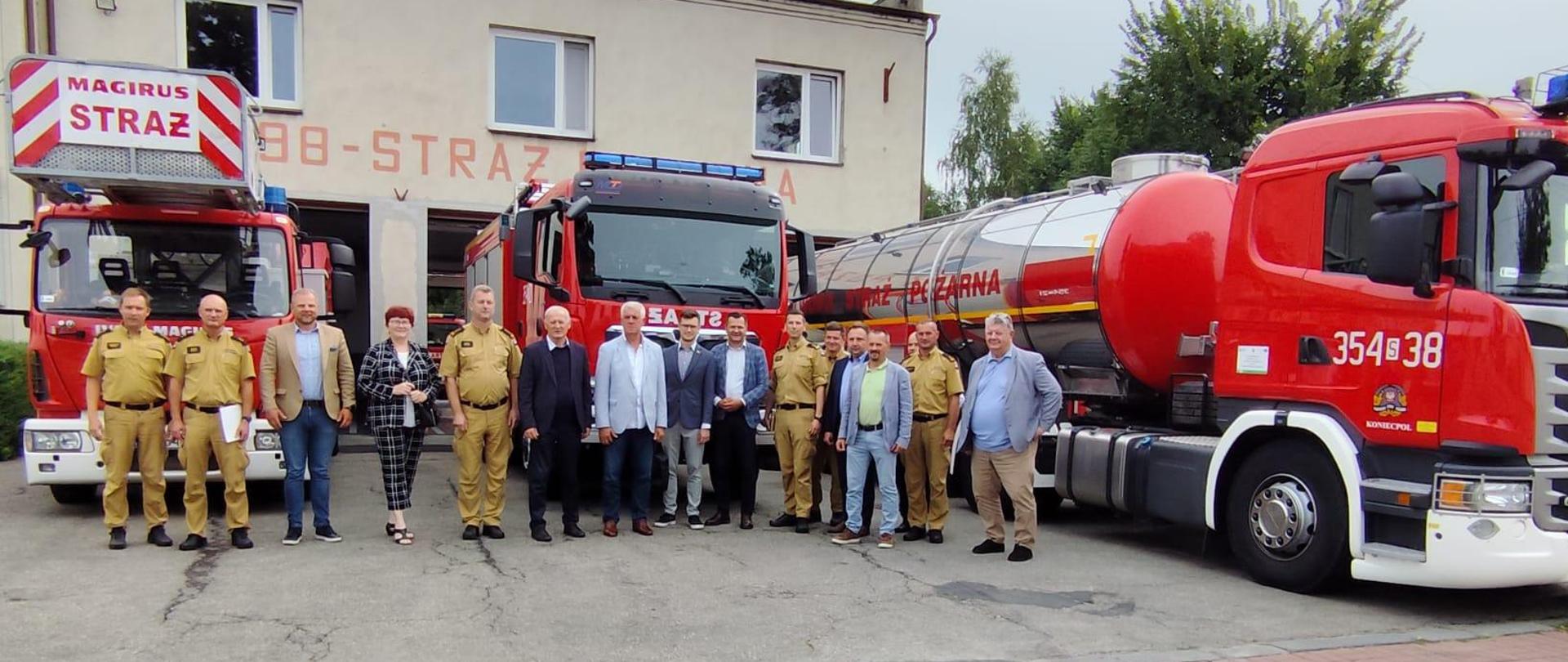 Sytuacja odbywa się przed budynkiem Jednostki Ratowniczo-Gaśniczej numer 4 w Koniecpolu. Przed bramy garażowe wyprowadzone zostały samochody strażackie. Przedstawiciele samorządów wraz ze strażakami pozują do zdjęcia grupowego na tle samochodów oraz budynku jednostki.