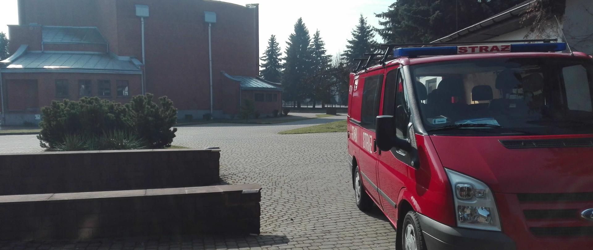 Zdjęcie przedstawia samochód strażacki, który stoi na placu obok kościoła. W tle widać murowany kościół.
