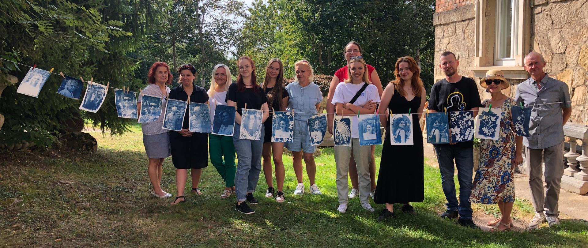 Fotografia przedstawia grupę osób w plenerze pomiędzy budynkiem po stronie prawej, a drzewem po stronie lewej. Na sznurku przed nimi powieszone są prace w formacie A4 w technice cyjanotypii 