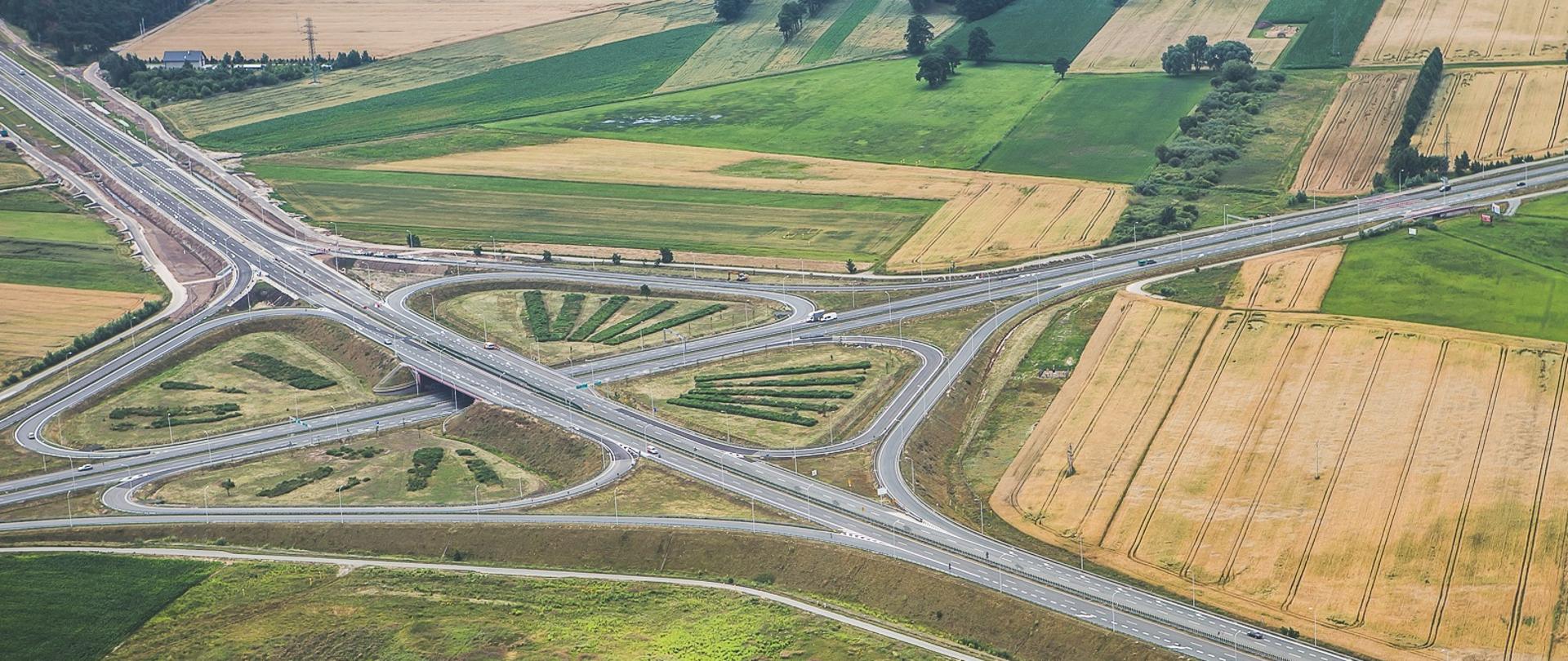 Zdjęcie pokazuje węzeł drogowy z lotu ptaka, w kształcie koniczyny, otoczone różnobarwnymi polami. Drogi łączą się i rozdzielają tworząc zakręty i zjazdy. Pola wokół są w różnych odcieniach zieleni i żółci, sugerując różne etapy wzrostu roślin. Na drogach widać kilka pojazdów.