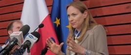 Minister funduszy funduszy i polityki regionalnej Katarzyna Pełczyńska-Nałęcz stoi przy mikrofonach, za jej plecami flagi Polski i UE